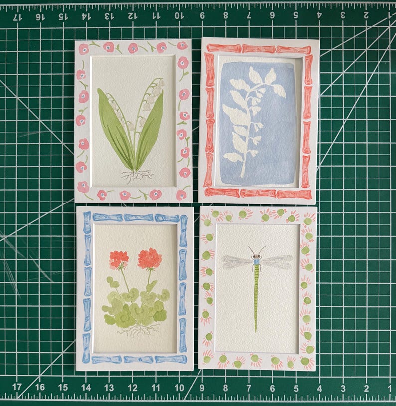 May include: Four rectangular white frames containing watercolor illustrations. Top left: lily of the valley with a pink floral border. Top right: white botanical silhouette on blue with a red bamboo-style border. Bottom left: orange flowers with a blue bamboo-style border. Bottom right: green dragonfly with a pink and green dotted border.