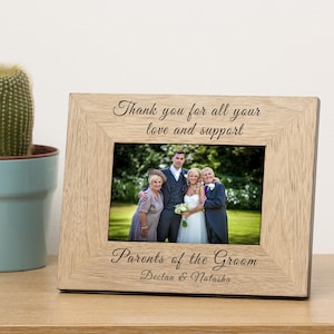 Pode incluir: Uma moldura de fotografia de madeira com acabamento marrom claro. A moldura tem um passe-partout branco e uma frente de vidro transparente. A moldura é gravada com o texto "Thank you for all your love and support" e "Parents of the Groom Declan & Natasha". A moldura contém uma foto de uma festa de casamento.