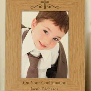May include: A light oak-colored picture frame with a cross design at the top. The frame holds a photo of a smiling child wearing a white shirt and patterned tie. The text "On Your Confirmation" and a name are engraved at the bottom.