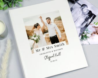 May include: A square white photo album featuring a centered wedding photograph of a couple holding hands on a beach. Below the image, the text reads Mr & Mrs Smith, Charlotte & Matthew, Stafford Hall, and the date 12.09.23 in black font.