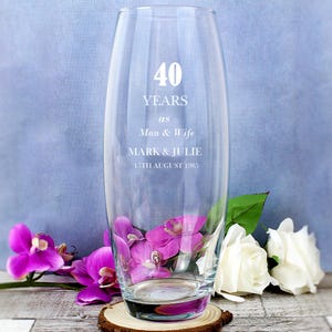 Peut inclure: Un vase en verre transparent avec le texte "40 YEARS as Man & Wife MARK & JULIE 15TH AUGUST 1985" gravé sur le côté. Le vase est décoré de fleurs roses et blanches.