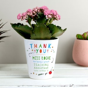 May include: White ceramic planter with a pink and green plant. The planter has a colorful design that says "Thank You" and "Miss Roche for being a great Teaching Assistant. Love Arthur x".