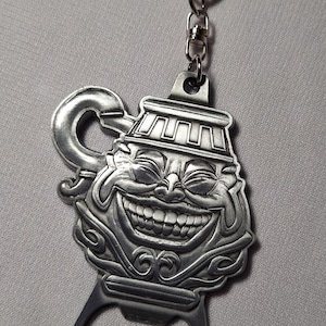 Puede incluir: Llavero abridor de botellas plateado con un diseño de cara sonriente. El llavero presenta una cara estilizada y detallada con una amplia sonrisa y un asa para abrir botellas. Incluye un llavero y un imperdible transparente.