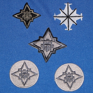 Pode incluir: Patches bordados em várias formas, incluindo estrelas e círculos, com um design geométrico detalhado em preto, branco e cinza. Os patches são colocados sobre um fundo de tecido azul vibrante.