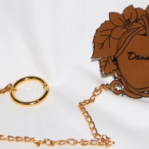 Könnte beinhalten: Eine goldene Kette mit einem Ring an einem Ende und einem braunen Metallanhänger mit einem stilisierten Apfel und dem Wort "Dandyism" am anderen Ende.