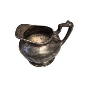 Peut inclure: Petite crémière antique en argent. Elle présente un corps arrondi, un bec verseur et une anse incurvée. La crémière a un aspect terni, suggérant l'âge et l'utilisation. Le design est simple et élégant, adapté pour servir de la crème ou du lait.