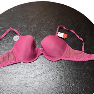 May include: A magenta-colored bra with adjustable straps and a lace trim along the bottom edge. The bra is displayed on a dark, textured surface. A tag is attached to the bra.