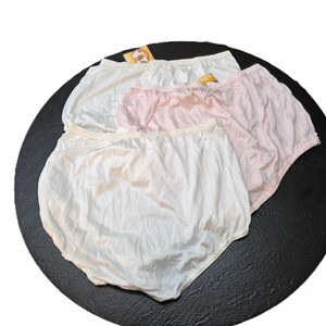 May include: Three pairs of underwear in white and pink are arranged on a black surface. The underwear has a high waist and a soft, silky appearance. Each pair has a tag attached.
