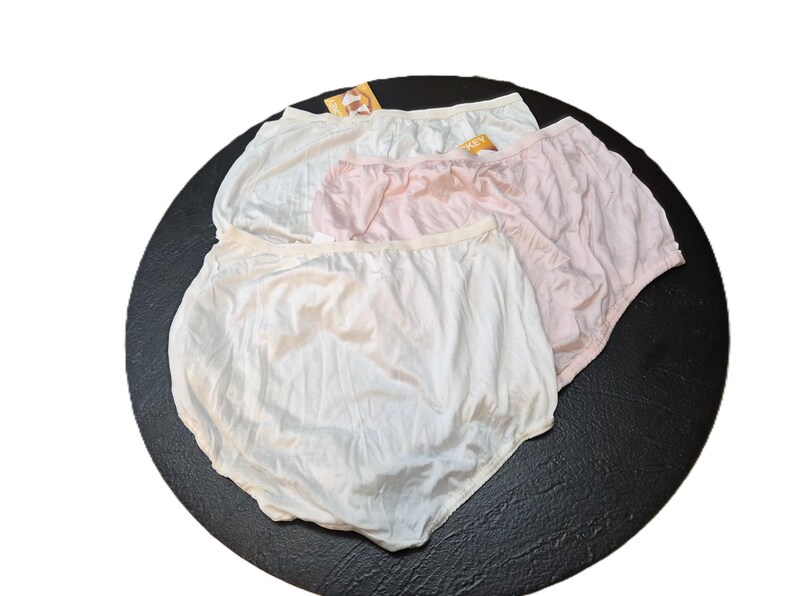 May include: Three pairs of underwear in white and pink are arranged on a black surface. The underwear has a high waist and a soft, silky appearance. Each pair has a tag attached.