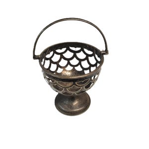 May include: A silver-coloured metal basket with a handle, featuring a scalloped design around the bowl. The basket sits on a small pedestal base. The handle is arched and attached to the rim.
