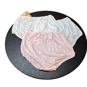 May include: Three pairs of underwear in white and pale pink are displayed on a black surface. The underwear has an elasticated waistband and a full coverage design. The fabric appears to be a soft, lightweight material.