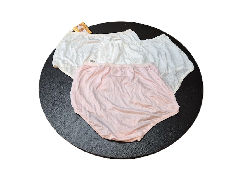 May include: Three pairs of underwear in white and pale pink are displayed on a black surface. The underwear has an elasticated waistband and a full coverage design. The fabric appears to be a soft, lightweight material.