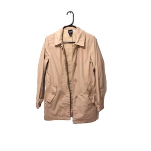 May include: A beige trench coat with a button-up front, collared neckline, and two front pockets. The coat is suspended from a black hanger. The brand name "GAP" is visible on the label.