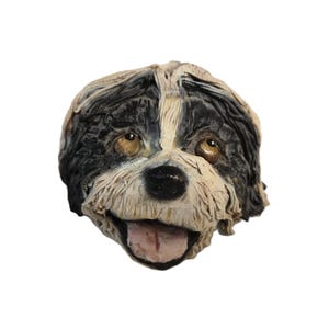 Könnte beinhalten: Eine detaillierte Skulptur eines Hundekopfes mit schwarz-weißem Fell, braunen Augen und einer rosa Zunge. Das Gesicht des Hundes ist mit strukturierten Details modelliert, was ihm ein lebensechtes Aussehen verleiht. Die Skulptur steht vor weißem Hintergrund.