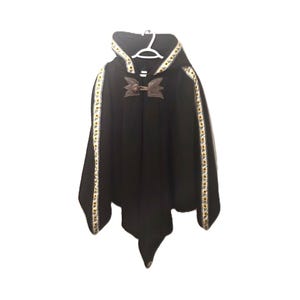 May include: A black hooded cloak featuring a decorative trim with a sunflower pattern along the hood and down the front panels. The garment is secured at the neck with a brown leather-like toggle closure and is displayed on a white plastic hanger.