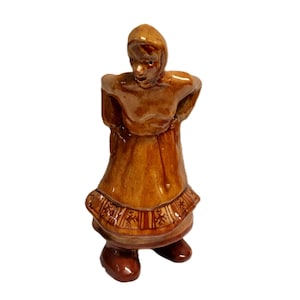 May include: A ceramic figurine of a woman in a traditional dress, with a head covering. The figurine is primarily brown with darker brown accents, and the dress features a decorative border. The figure's hands are positioned at her back.