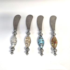 May include: Four butter knives with stainless steel blades and decorative glass handles. The handles feature various colors and patterns, including white with black stripes, beige with a leaf design, blue with black stripes, and gold with red accents.