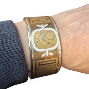 May include: A wide gold-tone metal cuff bracelet featuring a central white cartouche with an etched Egyptian-style figure. The surface displays a weathered, distressed finish with geometric border patterns flanking the central icon on both sides.