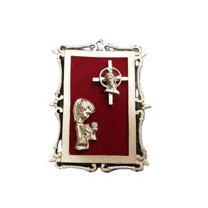 May include: Silver-toned religious wall hanging with a red velvet background. The frame has an ornate design. The image features a cross with a figure and a praying figure.