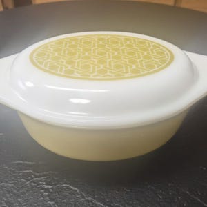 Op de afbeelding: Een vintage ovenschaal in geel en wit, met een geometrisch patroon op het deksel. De schaal heeft een afgeronde bodem en een deksel met een witte handgreep. Het deksel heeft een geel geometrisch ontwerp.