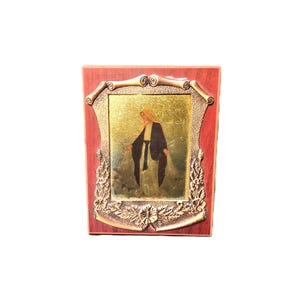 May include: A framed religious icon featuring a figure with a gold halo and dark robes. The image is set within a decorative gold frame, mounted on a red wooden base. The icon is a religious item.