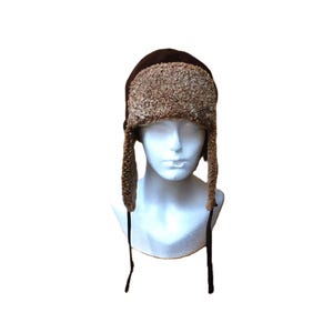 May include: A brown winter hat with ear flaps and a faux fur trim. The hat is displayed on a white mannequin head. The ear flaps have black strings that can be tied under the chin. The hat is designed to keep the head and ears warm in cold weather.