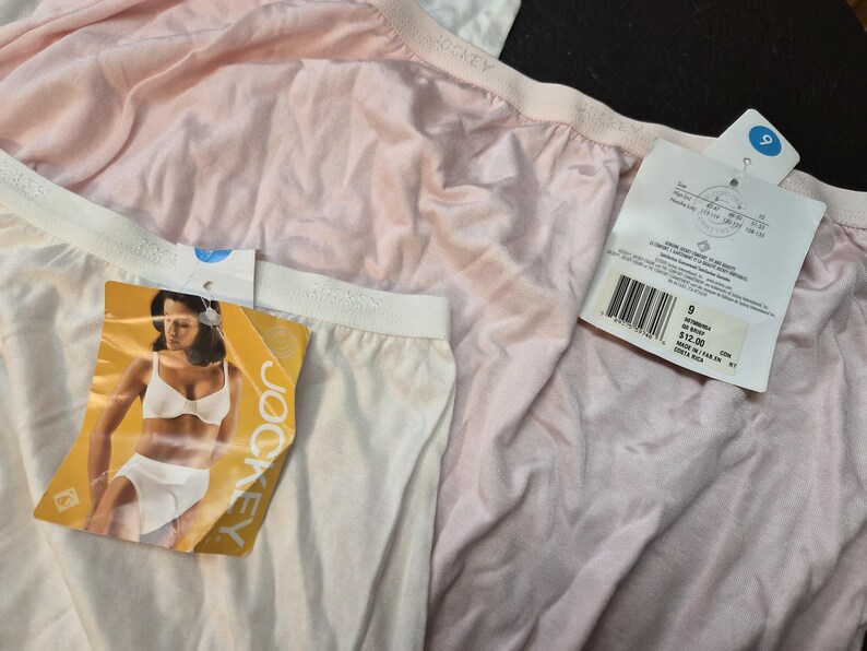May include: Close-up of three pairs of Jockey underwear in white and pale pink. Each pair has a white waistband and a tag attached. One tag features a woman in a bra and underwear. The other tags have product information.