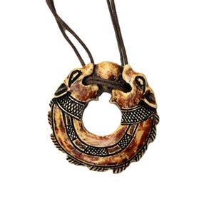 May include: A circular pendant necklace with a brown cord. The pendant is a light brown colour with darker brown accents and intricate carved details. The design appears to be of two animal heads facing each other.