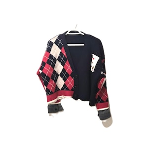 May include: A navy blue cardigan with a distinctive design. The left side has a red, black, and white argyle pattern, contrasting with the solid navy right side. Denim cuffs and a playing card symbol patch add detail.