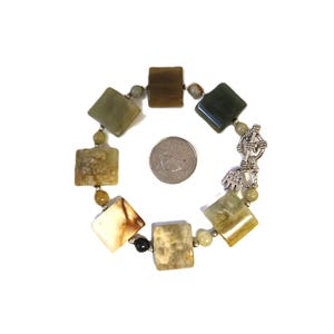Puede incluir: Una pulsera con piedras cuadradas verdes y beige, con pequeñas cuentas redondas. La pulsera tiene un cierre plateado y un dije. Las piedras miden aproximadamente 2 cm. La pulsera está sobre un fondo blanco.