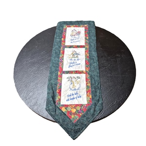 May include: A festive Christmas table runner with a green border and a central panel featuring three embroidered snowman scenes. Each scene has text: "There's no time like the presents", "Make a Joyful Noise", and "Deck the halls with boughs of holly".