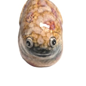 May include: A ceramic figurine shaped like a fish with a rounded body. The surface features a mottled pattern of tan, brown, and purple spots. It has two prominent, circular eyes with black pupils and a small, dark, horizontal mouth line.