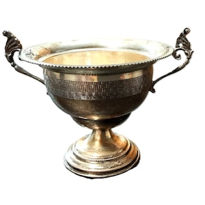 May include: A silver-toned metal trophy or decorative bowl with two ornate handles. The bowl has a wide rim and a textured band around the middle. It sits on a pedestal base.