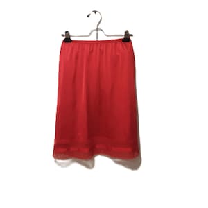 May include: A bright red slip skirt hanging on a metal hanger. The skirt is made of a smooth, shiny fabric and features a delicate lace trim along the hemline. The garment is displayed against a plain white background.