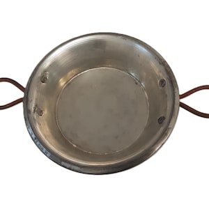 May include: A round, silver-colored metal pan with two brown, looped handles. The pan has a simple, utilitarian design, with a slightly textured surface and rivets securing the handles. Ideal for cooking.