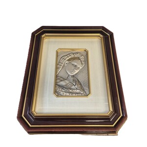 May include: A framed silver relief of a woman with a draped head covering. The artwork is set within a gold-toned border and a dark brown wooden frame. The frame has an octagonal shape.