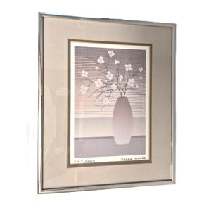 Könnte beinhalten: Gerahmter Kunstdruck mit einer Vase mit weißen Blumen und Zweigen. Das Kunstwerk ist vor einem Fenster mit horizontalen Jalousien platziert. Der Druck ist mit "Pierre Girard" signiert und enthält den Text "En Fleurs". Der Rahmen ist silberfarben.