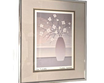 May include: Framed art print featuring a vase with white flowers and branches. The artwork is set against a window with horizontal blinds. The print is signed "Pierre Girard" and includes the text "En Fleurs". The frame is silver.