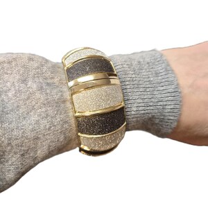 May include: A gold-coloured cuff bracelet featuring alternating bands of glittery silver and dark grey. The bracelet has a rectangular shape with rounded edges and is worn on a wrist. The bracelet is a fashion accessory.