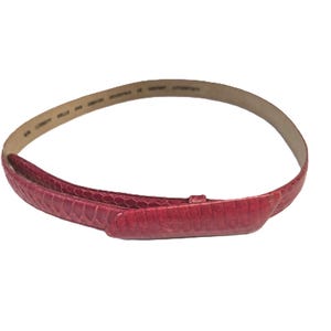 Op de afbeelding: Een rode riem met een textuur die lijkt op slangenleer. De riem heeft een rechthoekige gesp en een dunne, lichtgekleurde binnenvoering. De riem is gebogen.