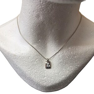 Op de afbeelding: Een zilveren ketting met een kleine, vierkante hanger met een heldere, rechthoekige edelsteen. De hanger hangt aan een fijne zilveren ketting, gepresenteerd op een witte mannequin buste. De ketting is eenvoudig en elegant.