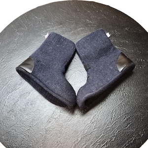 May include: A pair of navy blue baby boots with black leather detailing, set on a textured black surface. The boots are made of a soft, felt-like material and have a simple, classic design.