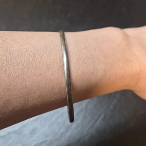 May include: A thin, smooth, metallic bangle bracelet with a polished silver-toned finish. The bracelet features a simple, continuous circular band design that rests against the skin, showcasing a clean and uniform appearance without any added embellishments.