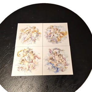 以下が含まれることがあります： パステルカラーのケルビムの姿を描いた、正方形のセラミックタイル4枚。各タイルには、翼のある複数のケルビムが描かれており、花や雲のような要素に囲まれています。タイルは黒い円形の表面に配置されています。