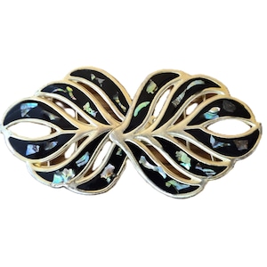 Puede incluir: Broche decorativo simétrico con marco de metal dorado. El broche presenta esmalte negro e incrustaciones iridiscentes de concha de abulón. El diseño recuerda hojas o alas estilizadas, creando un accesorio elegante.
