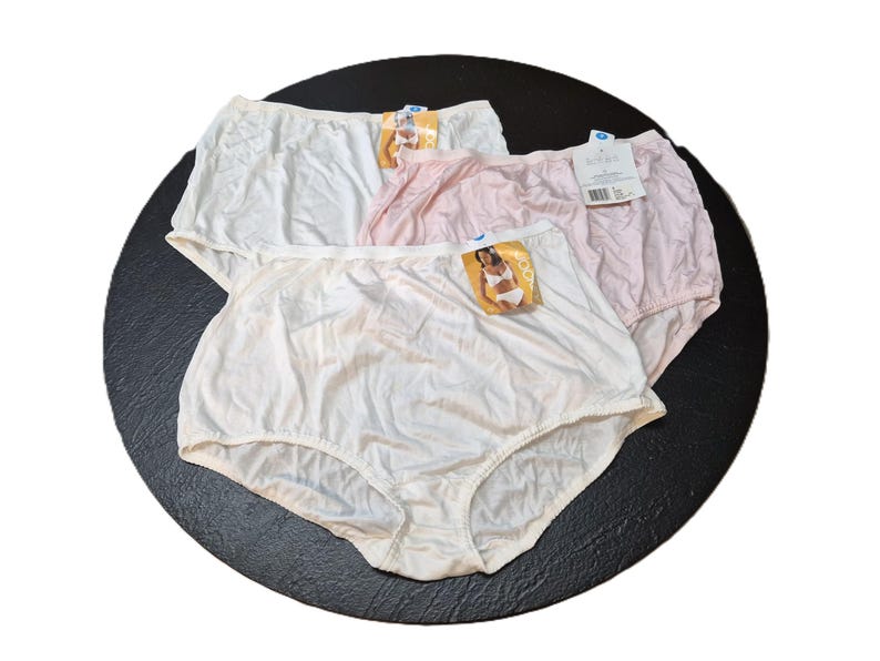 May include: Three pairs of high-waisted underwear in white and pink. Each pair has a tag attached. The underwear is made of a smooth, possibly satin-like material. The items are arranged on a dark, circular surface.