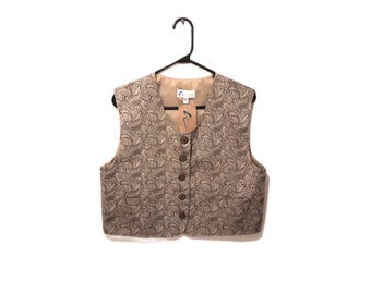 Puede incluir: Un chaleco beige con estampado de cachemira, abotonado y colgado de una percha negra. El chaleco tiene cuello en V y una etiqueta adjunta. El color neutro del chaleco lo hace adecuado para diversas ocasiones.