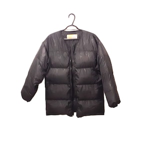 Puede incluir: Una chaqueta acolchada negra con cuello en V y cremallera completa. La chaqueta está colgada de una percha negra sobre un fondo blanco. Una etiqueta es visible en el escote.