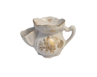 Puede incluir: Una taza de afeitar de porcelana blanca con un patrón floral dorado en el exterior y un borde festoneado con ribete de oro. La taza incluye un asa lateral y un compartimento integrado para la brocha, ambos con detalles en color oro.