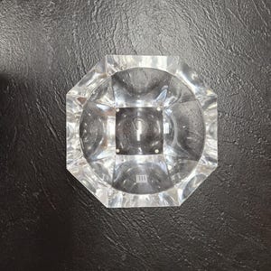 May include: A clear, octagonal ashtray with a circular interior. The ashtray is made of a transparent material, likely glass or crystal, and sits on a black surface. The design features geometric facets and a central square.